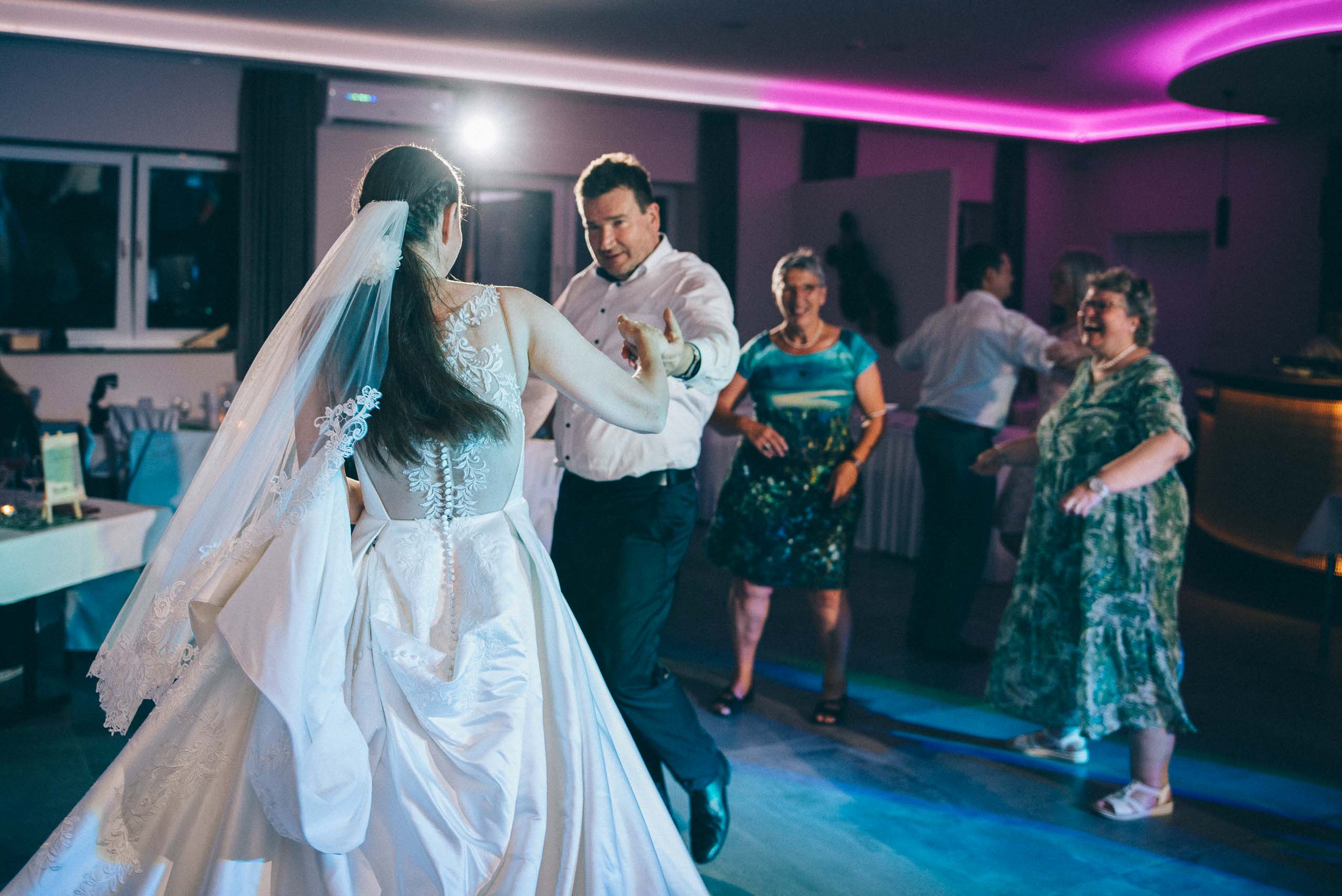 Szene einer Hochzeitsfeier. Der Hochzeitsfotograf hat das Brautpaar während des Hochzeitstanzes fotografiert. Das aufwändig gestaltete Hochzeitskleid ist von hinten zu sehen. Im Hintergrund tanzen Gäste.