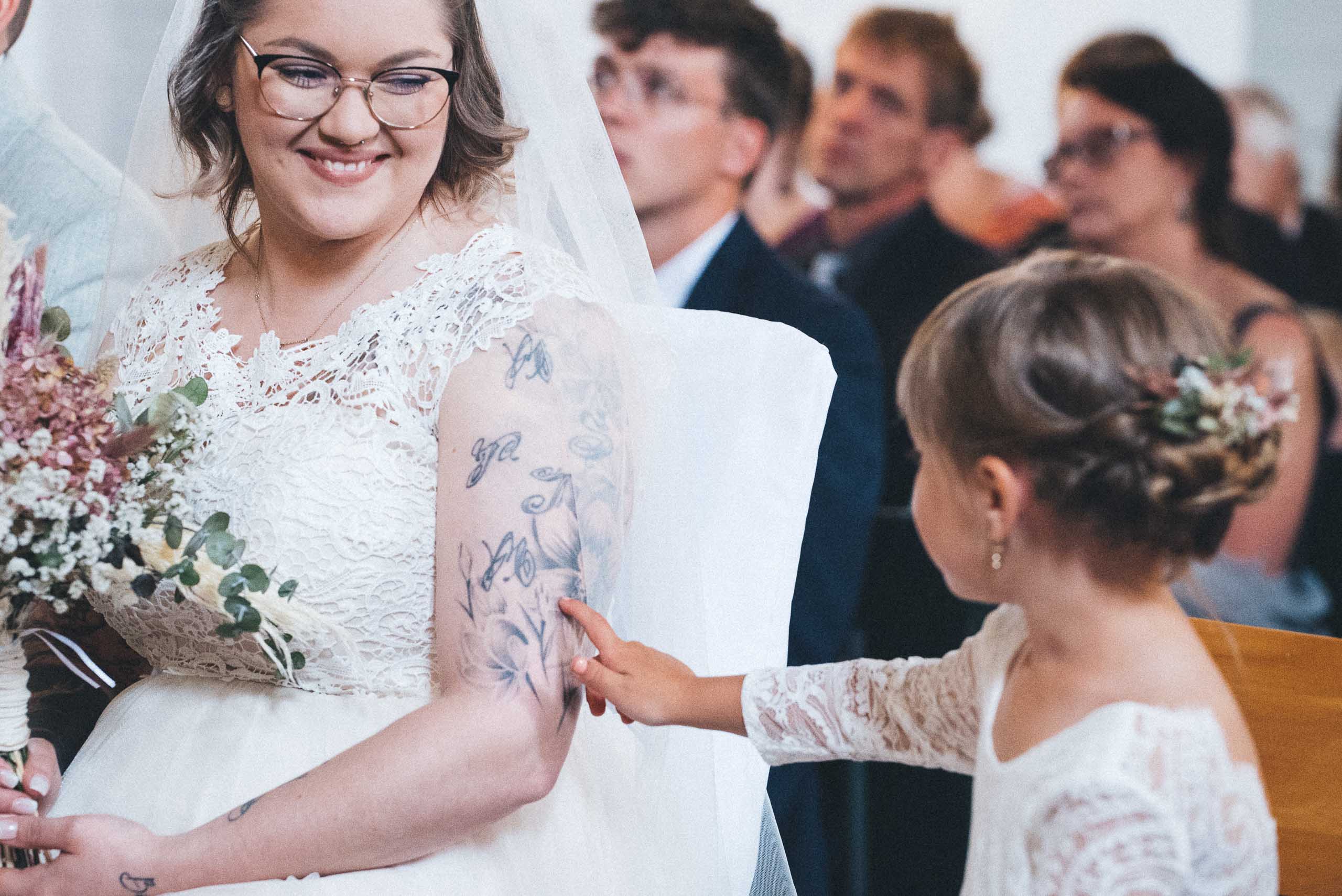 Momentaufnahme einer Hochzeit, die Braut im weißen Hochzeitskleid lächelt ihr Kind an. Das Kind berührt den Arm der Mutter.
